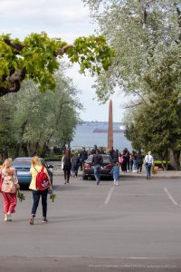 Фотография с улицы: люди прогуливаются по набережной у моря, на фоне виднеется памятник. Видна часть татуировки на руке одного из людей.