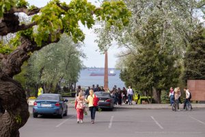 Фотография с видом на море: люди стоят у памятника, на фоне виднеется корабль. Видна часть татуировки на руке одного из людей.