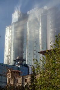 Фотография современного стеклянного небоскреба, освещенного яркими солнечными лучами, пробивающимися сквозь облака. Здание имеет строгие геометрические формы и отражающие поверхности.