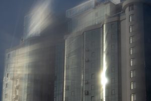 Фотография современного небоскреба, освещенного яркими солнечными лучами, пробивающимися сквозь облака. Здание имеет строгие геометрические формы и отражающие поверхности.