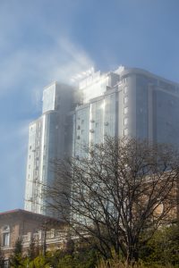 Фотография современного стеклянного небоскреба, освещенного яркими лучами солнца, пробивающимися сквозь облака. Архитектура здания отличается строгими линиями и отражающими поверхностями.