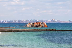 Фотография затонувшего корабля, расположенного у берега моря. На заднем плане видны городские постройки и голубое небо.
