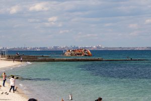 Фотография затонувшего корабля на фоне моря и городского пейзажа. Видны люди, отдыхающие на пляже.