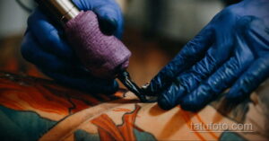 Фото тату процесса нанесения рисунка на кожу. Мастер в синих перчатках работает машинкой над татуировкой, вид крупным планом.