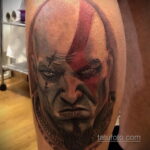 Фото тату с изображением Кратоса, персонажа из игры God of War, на плече. Реалистичный портрет с красным символом на лбу.