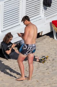 Фотография мужчины, стоящего на пляже в плавках и смотрящего в телефон. Видна татуировка на его ноге. Яркий солнечный день.