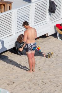 Фотография мужчины, стоящего на пляже в плавках и демонстрирующего татуировку на ноге. Яркий солнечный день, пляжные зонтики на заднем плане.