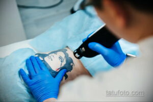 Фотография процесса нанесения портретной татуировки на ноге клиента: мастер работает тату-машинкой, используя голубые перчатки.