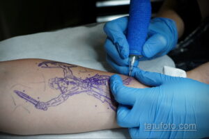 Фото тату процесса переноса эскиза татуировки на кожу. Мастер в голубых перчатках наносит контуры рисунка на руку клиента.
