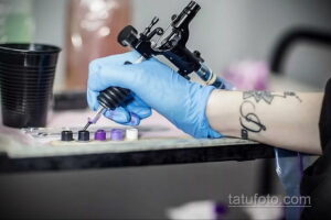 Фото тату процесса нанесения татуировки: рука мастера в голубых перчатках работает с тату-машинкой и красками.