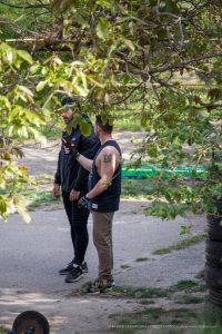 Фото тату: мужчина с татуировкой иероглифов на плече, стоящий на улице под деревом. Изображение передает атмосферу свободы и уличного стиля.