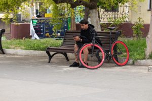 Фотография молодого человека, сидящего на скамейке с велосипедом. Акцент на татуировках на ногах и общем стиле.