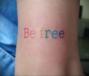 Фото татуировки с надписью 'Be free' на запястье, выполненной в радужных цветах. Мотивирующая фраза, яркие цвета.