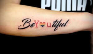 Фото тату с надписью 'Be You-tiful', где слово 'You' заменено на радужное сердце, выполненное в каллиграфическом стиле.