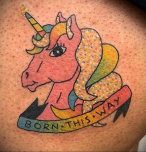 Фото тату единорога с радужной гривой и надписью 'Born This Way' на ленте, выполненное в ярких и красочных тонах.