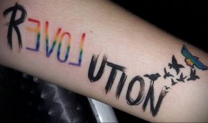 Фото тату надписи 'REVOLUTION', выполненной в цветах радуги, с летящими птицами. Символичный и яркий рисунок татуировки, выражающий протест и стремление к переменам.