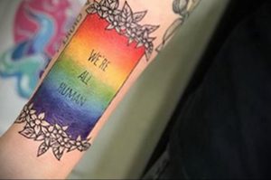 Фото тату: радужная полоса с надписью "We're All Human" (Мы все люди), обрамленная цветами. Символичный эскиз, выражающий поддержку равенства.