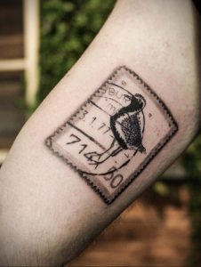 Фото тату на руке, изображающее птицу в рамке почтовой марки. Черно-белый рисунок татуировки в минималистичном стиле.