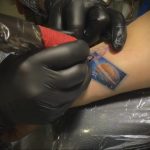 Фото процесса татуировки: мастер в черных перчатках наносит рисунок татуировки в виде марки с изображением планеты на руку клиента.