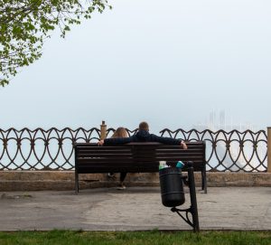 Фотография пары, сидящей на деревянной скамейке у моря в туманный день. Вид сзади, акцент на спокойной атмосфере и морском пейзаже.