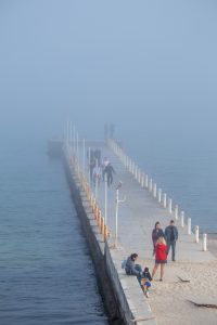 Фотография людей, идущих по пирсу в тумане. Пирс окружен водой, а вдалеке виднеются силуэты других людей.