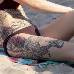 Крупный план татуировки на боку девушки, изображающей дракона, обвивающегося вокруг ярких красных цветов. Детализированный рисунок татуировки.