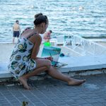 Фотография женщины, рисующей пейзаж на холсте на фоне моря и пляжа. Художница сидит на парапете с палитрой и кистями.