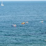 Фотография моря с парусной яхтой, каяками и людьми, плавающими в воде. Голубое небо и спокойное море создают летнюю атмосферу.