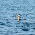 Фотография мужчины, стоящего в море с удочкой, на фоне спокойной воды и голубого неба.