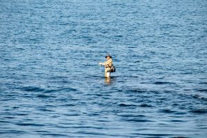 Фотография мужчины, стоящего в море с удочкой, на фоне спокойной воды и голубого неба.