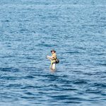 Фотография мужчины, стоящего в море с удочкой, на фоне спокойной воды и голубого неба.