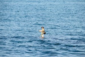 Фотография мужчины, стоящего в море с удочкой, на фоне спокойной воды и голубого неба.