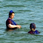Фотография двух женщин, плавающих в море, одна смотрит в сторону, другая повернулась лицом к зрителю.