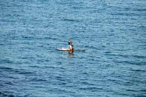 Человек, стоящий на доске для SUP-серфинга в синем море, солнечный день, спокойная вода, вид с расстояния.