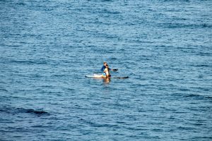 Человек, стоящий на доске для SUP-серфинга в синем море, солнечный день, спокойная вода, вид с расстояния.