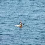 Два человека на доске для SUP-серфинга в синем море, солнечный день, спокойная вода, вид с расстояния.