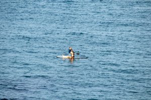 Два человека на доске для SUP-серфинга в синем море, солнечный день, спокойная вода, вид с расстояния.