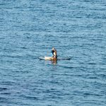 Человек, стоящий на доске для SUP-серфинга в синем море, солнечный день, спокойная вода, вид с расстояния.