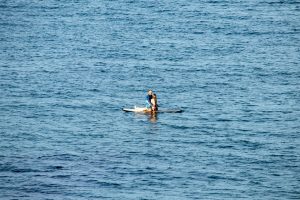 Человек, стоящий на доске для SUP-серфинга в синем море, солнечный день, спокойная вода, вид с расстояния.