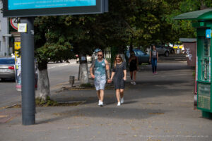 Фотография пары, идущей по улице, держась за руки. У мужчины видны татуировки на руках, а на фоне городская улица и деревья.