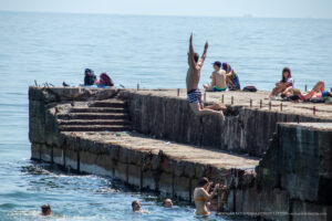 Фотография с пляжа, где парень прыгает в воду с бетонного пирса, окруженный отдыхающими. Видны яркие купальники и голубое море.
