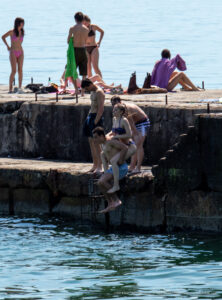 Фотография с пляжа, где молодой человек прыгает в воду с бетонного пирса, окруженный отдыхающими. Видны яркие купальники и солнечный свет.