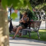 Фото мужчины, сидящего на скамейке и говорящего по телефону. На его руке видна татуировка, частично скрытая одеждой. Фон размытый, с деревьями и транспортом.