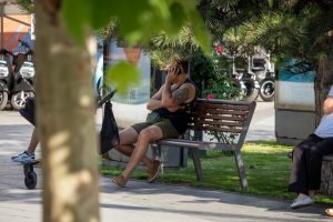 Фото мужчины, сидящего на скамейке и говорящего по телефону. На его руке видна татуировка, частично скрытая одеждой. Фон размытый, с деревьями и транспортом.