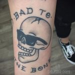 Фото тату: череп в солнцезащитных очках с надписью "BAD TO THE BONE". Классический стиль традишн с четкими линиями и яркими цветами.