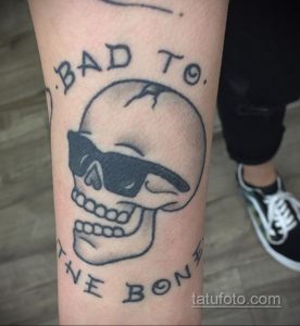 Фото тату: череп в солнцезащитных очках с надписью "BAD TO THE BONE". Классический стиль традишн с четкими линиями и яркими цветами.
