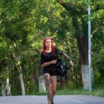 Фото девушки с красными волосами и татуировкой на ноге, идущей по улице в летний день.