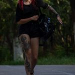 Фото девушки с красными волосами и татуировкой на ноге, идущей по улице в летний день.