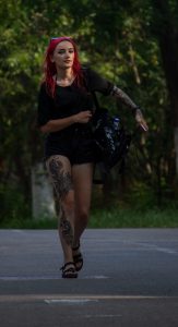 Фото девушки с красными волосами и татуировкой на ноге, идущей по улице в летний день.