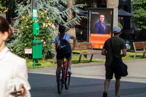 Фотография девушки, едущей на велосипеде по городу. На ее предплечье видна минималистичная татуировка из трех черных линий.